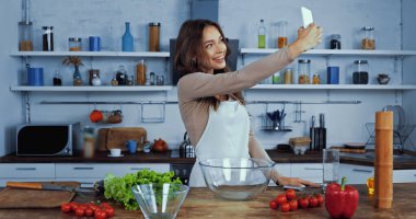Önlüklü neşeli kadın masadaki malzemelerin yanında selfie çekiyor.