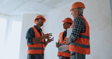 Builder gesturing while talking with coworkers in hard hats on construction site clipart