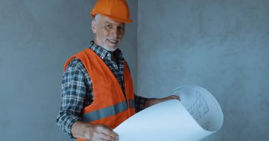 happy builder holding blueprint and smiling on construction site clipart