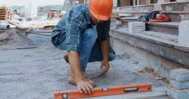 Builder in hard hat measuring ground with building level on construction site clipart