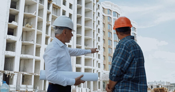 Engineer with blueprints pointing with hand while talking with builder on construction site