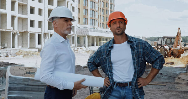 mature engineer with blueprints talking with builder on construction site