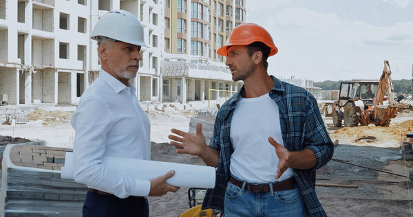 Engineer with blueprints talking with builder in hard hat on construction site