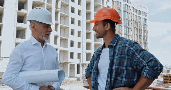 Engineer with blueprints talking with happy builder on construction site
