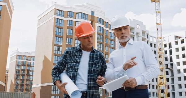 mature engineer and builder with gadget and blueprint talking on construction site