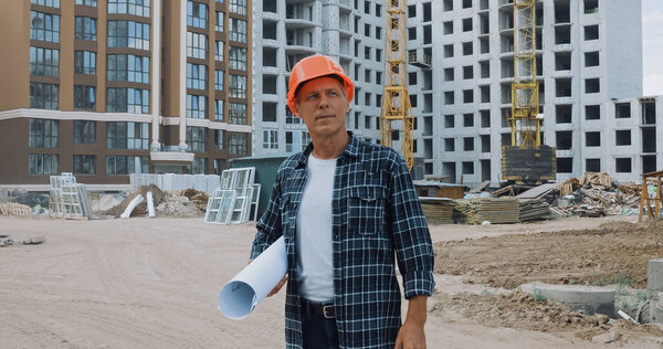 mature builder holding blueprint and standing on construction site