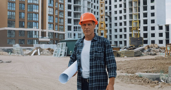 middle aged builder holding blueprint and standing on construction site
