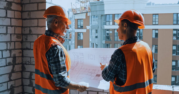 Builders in safety vests with blueprint talking on construction site