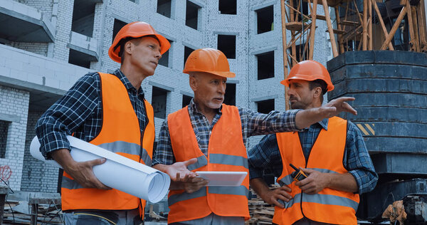 mature worker pointing with hand and talking near coworkers and building crane on construction site
