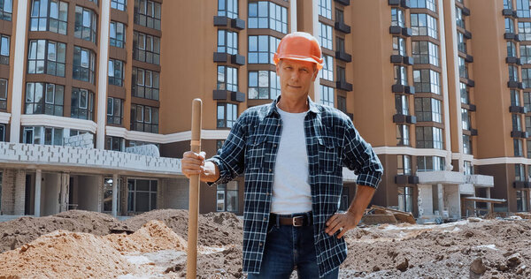 Smiling builder holding shovel and standing with hand on hip on construction site