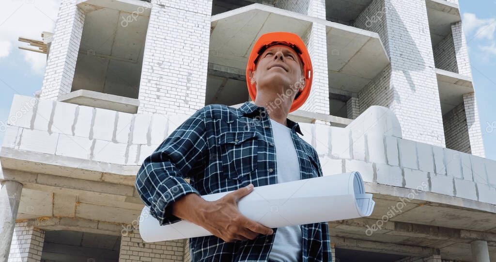 low angle view of mature builder holding blueprint and smiling on construction site