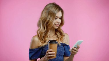 Sarışın kadın akıllı telefon kullanıyor ve pembe üzerinde izole olmak için kahve tutuyor.
