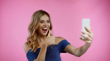 Mutlu genç bir kadın pembe üzerine izole edilmiş selfie çekerken baş parmağını gösteriyor. 