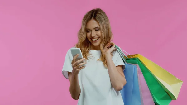 Akıllı telefon kullanan mutlu bir kadın ve mor renkte izole edilmiş alışveriş torbaları tutan.
