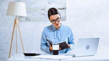 Yoğunlaştırılmış iş kadını kahve ve dizüstü bilgisayarın yanındaki deftere bakıyor.