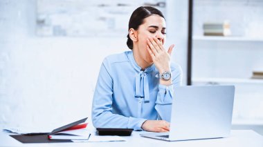 Yorgun iş kadını dizüstü bilgisayarın yanında esnerken ağzını kapatıyor.