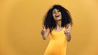Cheerful pregnant hispanic woman showing thumbs up isolated on yellow clipart