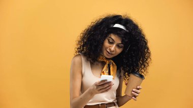 Gülümseyen İspanyol kadın akıllı telefon kullanıyor ve sarıda izole olmak için kahve tutuyor.