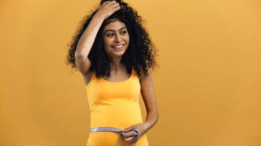 Happy pregnant hispanic woman holding tape measure isolated on yellow clipart