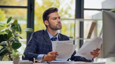 Kağıtları tutarken bilgisayar ekranına bakan sakallı iş adamı 