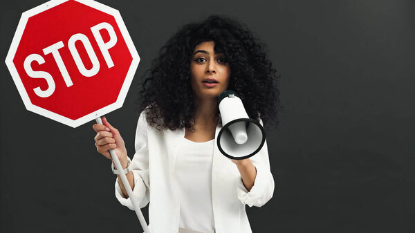 hispanic woman holding stop signboard and loudspeaker isolated on black 