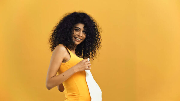 Счастливая беременная латиноамериканка держит детскую одежду изолированной на желтый