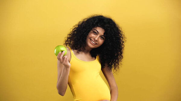 Беременная латиноамериканка с яблоком, улыбающаяся в камеру, изолированную на желтом