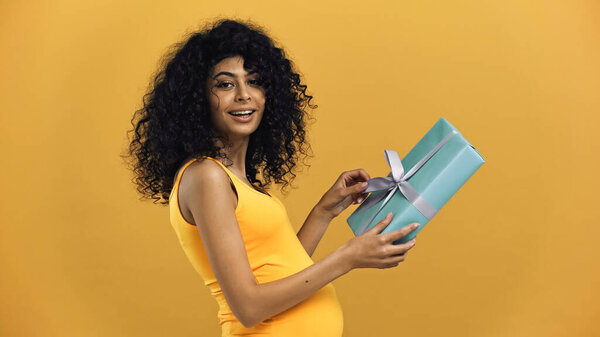Положительная беременная латиноамериканка держит подарочную коробку изолированную на желтый
