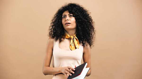 Страшная латиноамериканка, держащая блокнот изолированным на бежевом 