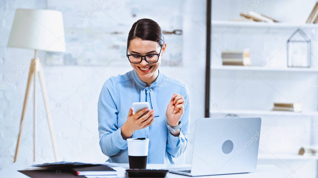 joyful businesswoman using smartphone near coffee to go and laptop at workplace
