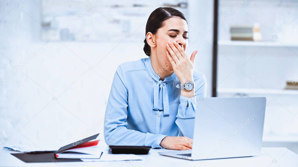 tired businesswoman covering mouth while yawning near laptop