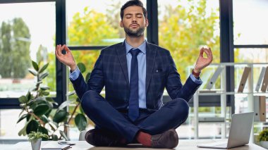businessman in suit meditating on desk in office  clipart