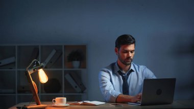 Takım elbiseli işadamı geç saatlere kadar çalışırken dizüstü bilgisayar kullanıyor.