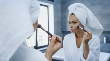young woman applying face powder with cosmetic brush while looking at mirror in bathroom clipart