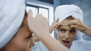 displeased young woman squeezing pimple and looking at mirror in bathroom clipart