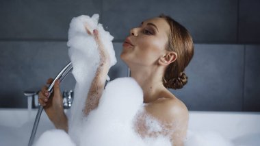 Genç bir kadın küvette banyo yapıyor ve beyaz köpük üflüyor. 