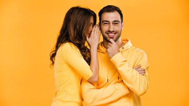 young woman whispering in ear of happy boyfriend isolated on yellow  clipart