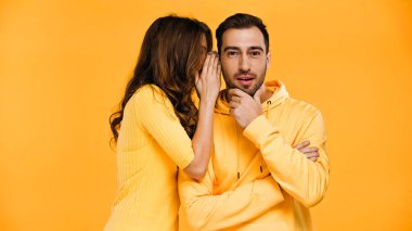 young woman whispering in ear of boyfriend isolated on yellow  clipart