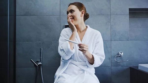 happy woman in bathrobe holding pregnancy test in bathroom