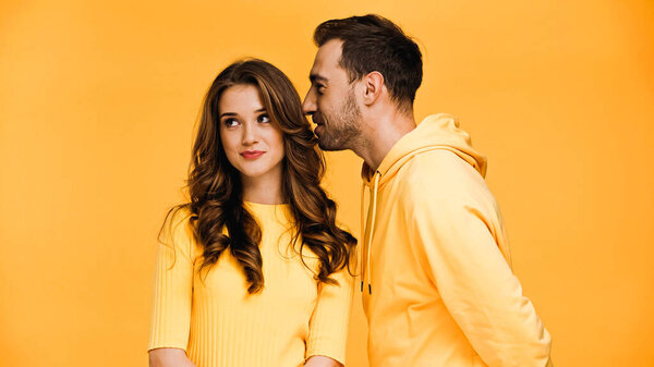 boyfriend whispering in ear of happy girlfriend isolated on yellow 