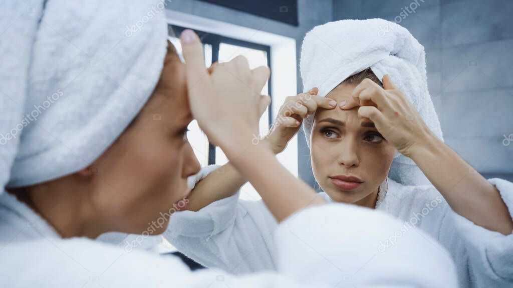 worried young woman squeezing pimple and looking at mirror in bathroom