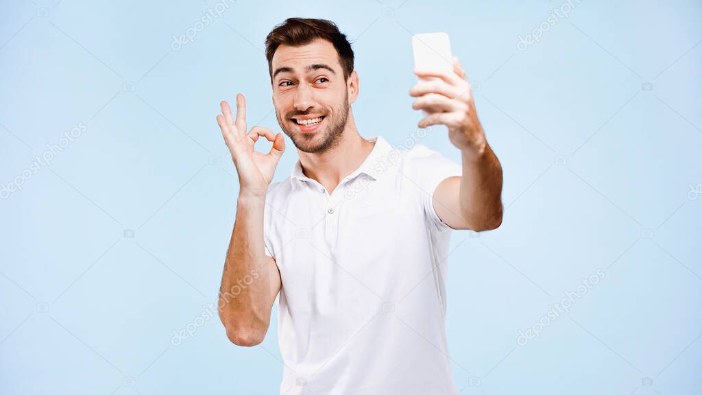 smiling man taking selfie and showing okay sign isolated on blue 