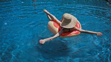 Hasır şapkalı bir kadının yüzme yüzüğüyle havuzda yüzüşünün arka görüntüsü.