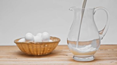 milk pouring into glass jar near wicker basket with white eggs on wooden table isolated on grey clipart