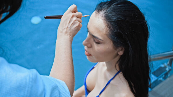 высокий угол зрения визажиста нанося порошок для лица с косметической щеткой на брюнетку женщина около бассейна 