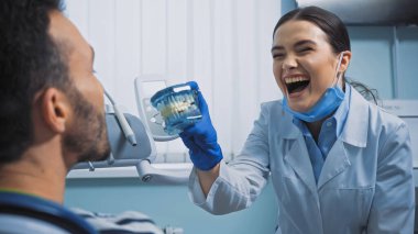 cheerful dentist showing jaw model to man in dental clinic clipart