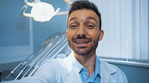 cheerful dentist looking at camera near lamp and dental equipment on blurred background
