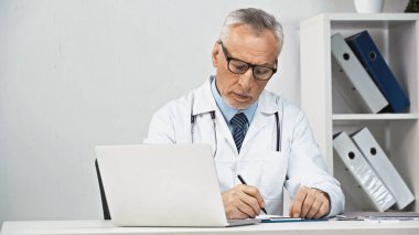 Ciddi bir doktor iş yerindeki dizüstü bilgisayarın yanındaki panoya teşhis koyuyor.