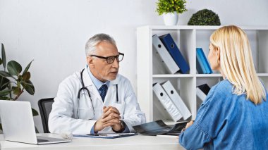 Gözlüklü olgun bir doktor danışma odasında sarışın bir kadınla konuşuyor.