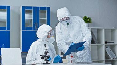 Tehlikeli madde giysisi içindeki bilim adamı laboratuardaki meslektaşlarına araştırma sonuçlarını gösteriyor.